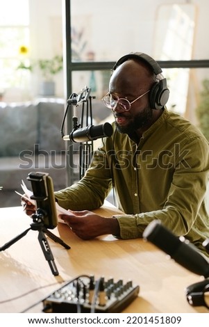 Young black man in headphones talking in microphone while reading his notes or plan of discussion in front of smartphone camera