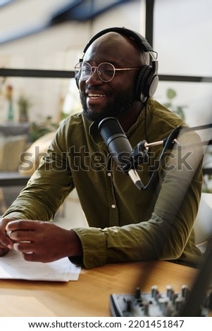 Young cheerful black man in headphones speculating on topical subject or curious news while sitting by desk and talking in microphone