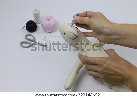 Tailoring of handmade textile toys for home and interior. Embroider black eyes on the rabbit with a needle. Hand sewing process with cloth, scissors, sewing accessories.
