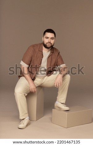 full length shot of attractive Hispanic man in his 30s sitting on blocks and posing confidently on neutral background