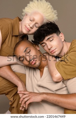 Portrait of a non-binary caucasian person in their 20's, a Black woman with albinism in her 20's, and a Hispanic man in his 20's with leaning against each other on a neutral background. Royalty-Free Stock Photo #2201467449