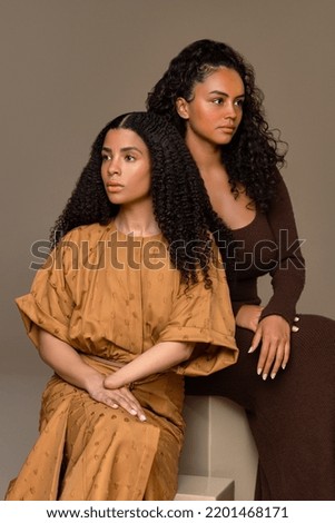 Studio portrait of an Afro-Indigenous woman in her 20's and an Afro-Latina in her 30's looking in opposite directions on a neutral background.