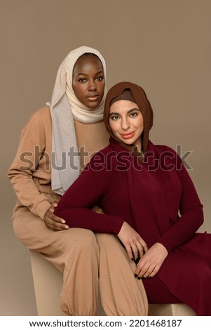 Studio portrait of an Afro-Arab woman in her 20's and an Arab Muslim woman in her 20's smiling together on a neutral background.