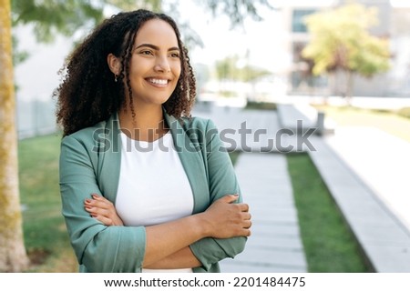 Portrait of lovely positive confident successful brazilian or latino woman with curly hair, business lady, in stylish elegant clothes, stands outdoors with arms crossed, looking ahead, smiles friendly Royalty-Free Stock Photo #2201484475