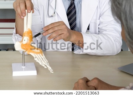 Orthopedic Surgeon in white gown and stethoscope pointing at human skeleton foot ankle bone joint anatomy model, present to the elderly patient about treatment of the ankle with surgery. Royalty-Free Stock Photo #2201513063