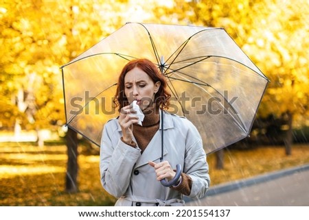 Unhappy caucasian millennial red-haired woman in raincoat with umbrella walks in rain coughs in napkin in park in autumn with yellow leaves. Health problems, cold and flu during covid-19 quarantine Royalty-Free Stock Photo #2201554137