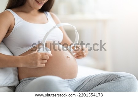 Cropped of expecting lady in comfortable homewear sitting on bed at home, holding white wireless headphones on her big belly, copy space. Music and baby development during pregnancy concept