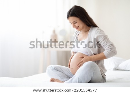 Smiling young pregnant woman sitting on bed in white bedroom at home, hugging her big tummy, feeling joyful as emotional well affect to baby neurological and psychological development, empty space Royalty-Free Stock Photo #2201554577