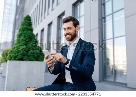 Businessman at conference outdoor. Happy confident attractive young caucasian male with beard in suit typing on smartphone near modern office building. App for successful business in city, new normal Royalty-Free Stock Photo #2201554771