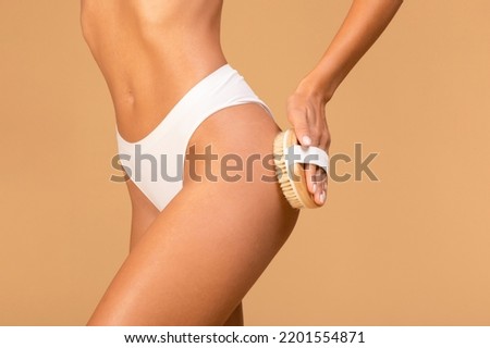 Young slim woman making anti cellulite or lymphatic drainage thigh massage, lady dry brushing her legs, exfoliating skin, taking care of her body, beige studio background