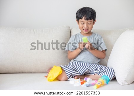 Adorable happy Asian little boy wearing a gray shirt and blue-white striped shorts sitting on a cream sofa is smiling happily is choosing his toys intentionally.