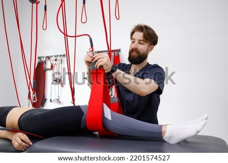 Physiotherapist assists female patient undergoing rehabilitative physiotherapy on a suspension rope system. Modern clinic using advanced rehabilitating therapy