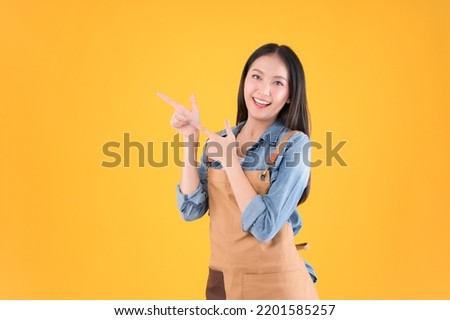 entrepreneur pretty asian female smile wearing apron freelancer own shop show and present pointing fingers copy space for your advertisement yellow background.