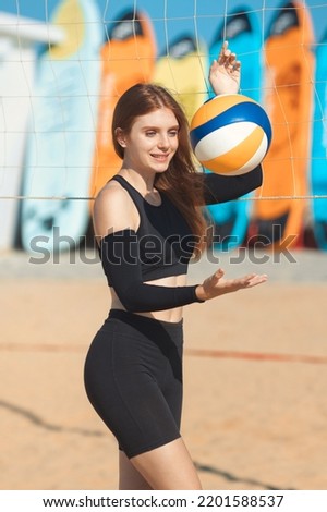 Happy beautiful ginger girl playing with volleyball ball at beach against the background of colored surfboards on summer sunny day 