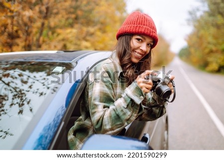 Beautiful woman takes pictures on the camerafrom the car window. Smiling woman enjoying autumn weather. Rest, relaxation, lifestyle concept.