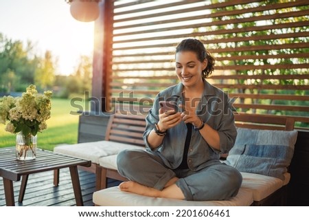 Happy young woman is using a phone sitting on the patio in summer. 