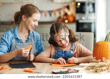 Happy smiling family mother and daughter making Halloween home decorations together while sitting at wooden table, mom and girl painting pumpkins and making paper cuttings