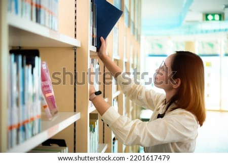 woman stands in library, looking for a book. asian woman in eyeglasses searching information for a project and working