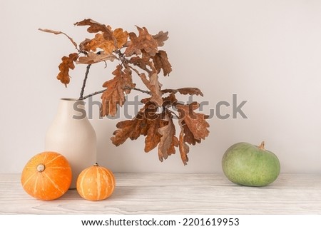 Autumn season concept. Dry oak leaves in vase with decorative orange and green pumpkins on light wooden table. Royalty-Free Stock Photo #2201619953