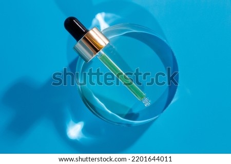 Glass pipette of hyaluronic acid on glass circle support, blue background. Cosmetics concept
