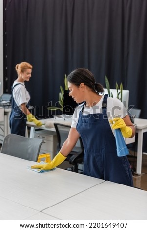woman with detergent and rag cleaning office desk near colleague on blurred background