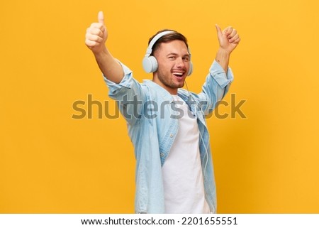 Dancing happy tanned handsome man in blue basic t-shirt headphones show thumbs up gesture Like posing isolated on orange yellow studio background. Copy space Banner Mockup. Time to relax concept