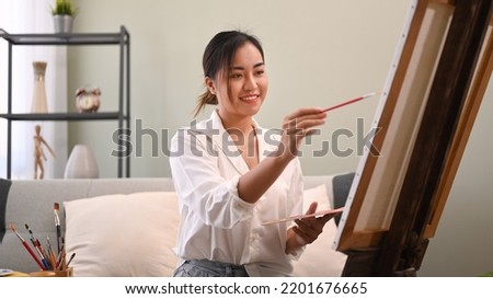Smiling female artist painting with watercolor on canvas in bright living room. Art, creative hobby and leisure activity concept