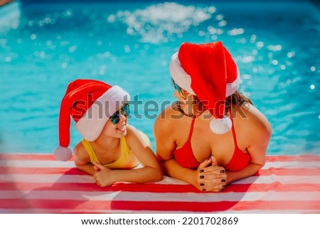 Beautiful young mother in red bikini and Christmas cap posing with cheerful daughter near the swimming pool. New Year celebration concept. Focus is at the woman. Royalty-Free Stock Photo #2201702869