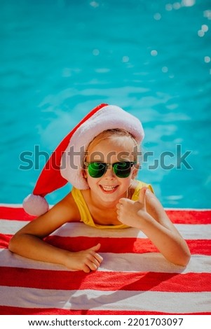 Cute young girl in yellow swim suite with Christmas cap and sunglasses posing near the swimming pool on red and white stripped towel. New Year celebration concept. Royalty-Free Stock Photo #2201703097