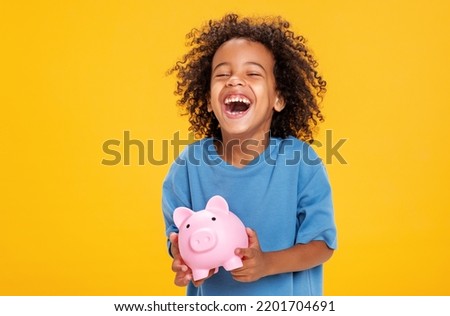 Positive african american boy in casual outfit laughing and saving money in piggy bank against yellow background Royalty-Free Stock Photo #2201704691