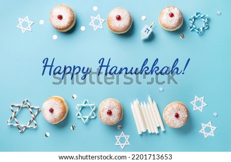 Hanukkah sweet doughnuts sufganiyot (traditional donuts) with fruit jelly jam, gift boxes, spinnig driedel and candles on blue paper background. Jewish holiday Hanukkah concept. Top view, copy space.