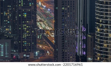 Highway and overpass between skyscrapers with glowing windows in Dubai downtown aerial night timelapse. Glowing windows in towers near metro station and busy traffic from above.
