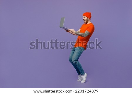 Full body young surprised IT man he wear red hat t-shirt hold use work on laptop pc computer stand on toes leaning back isolated on plain pastel light purple background studio People lifestyle concept
