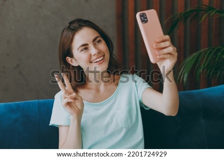 Young fun smiling woman she 20s in casual clothes mint t-shirt do selfie shot on mobile cell phone post photo on social network show v-sign sit on blue sofa indoor rest at home in own room apartment