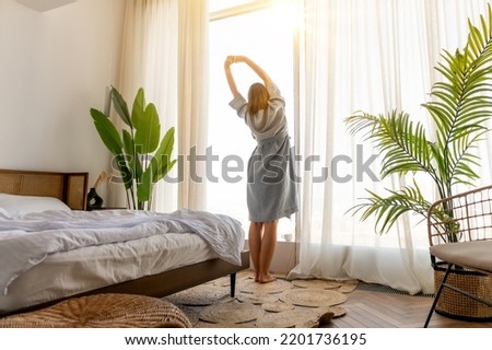 Rear view woman sitting on bed in bedroom, stretching hands after awakening, dreamy young female enjoying morning, starting new day in modern apartment with large panoramic windows Royalty-Free Stock Photo #2201736195