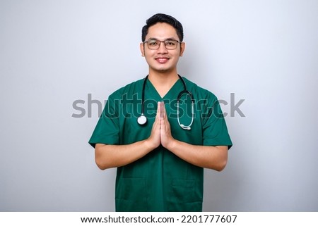 Smiling beautiful asian male doctor, physician in scrubs smiling, hold hands together over chest in namaste, greeting gesture isolated white background