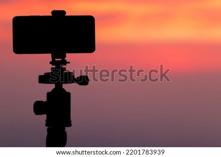 Silhouette of smartphone mounted on tripod to capture time-lapse video of the beautiful colors in the sky during twilight. photographer uses smartphone to record video of sky backdrop during Twilight.