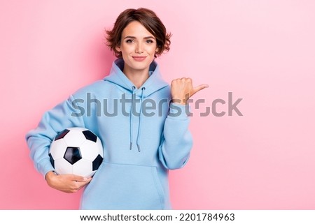 Photo of young adorable gorgeous nice pretty girl wear blue hoodie hold football ball finger pointing empty space positive recommend isolated on pink color background