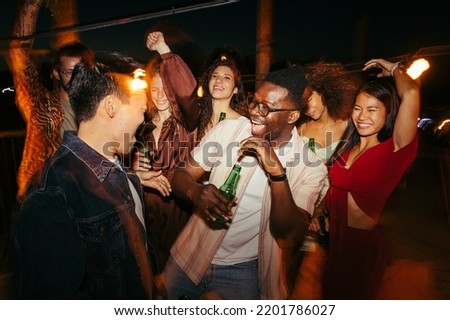 A cheerful group of multiracial people is dancing, drinking beer, and having a good time at the outdoor nighttime event.