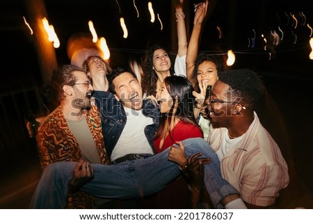 The interracial friends are carrying a man and tossing him in the air at the rooftop night summertime outdoor party. They are having a good time, dancing and drinking beer. Royalty-Free Stock Photo #2201786037