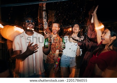 A cheerful group of multiracial friends is celebrating and having fun with confetti at the open-air summer rooftop nightclub. They are having a blast, dancing and drinking beer. Royalty-Free Stock Photo #2201786055