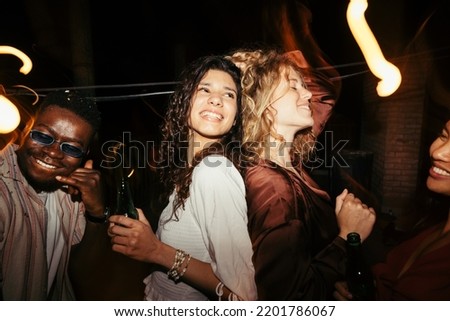 Playful girls dance at the summertime rooftop party at night. They are drinking beer and having a blast with their friends. Nightlife in capital cities. Royalty-Free Stock Photo #2201786067