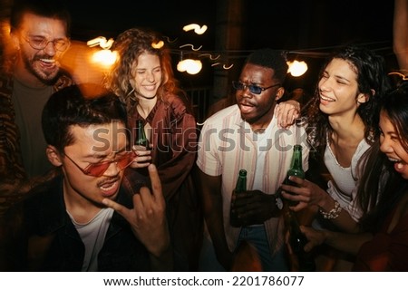 A multiracial group of friends is drinking beer and having a good time at the rooftop night party. Open-air clubbing concept. Royalty-Free Stock Photo #2201786077