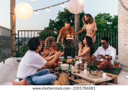 A group of interracial friends celebrates their friend's birthday at the rooftop party. A man is bringing the cake while the rest of the crew is clapping and singing a birthday song.