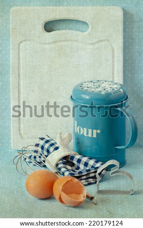 Old white kitchen cutting board with copy-space for writing menu, flour strainer, napkin and napkin ring, whisk and eggs. Toned photo. Vintage style.
