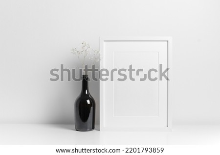 Blank frame mockup in white room with dark bottle wih flower decorations