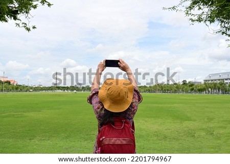 A female tourist raises a mobile phone to take pictures of the beauty of the green lawn. The blue sky and white clouds were scattered. Woman wearing yellow wide-brimmed hat and carrying red backpack.