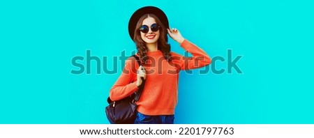 Portrait of happy smiling young woman wearing black round hat, sweater, backpack on blue background