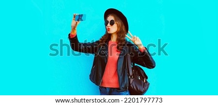 Portrait of young woman taking selfie with smartphone wearing black round hat, backpack on blue background