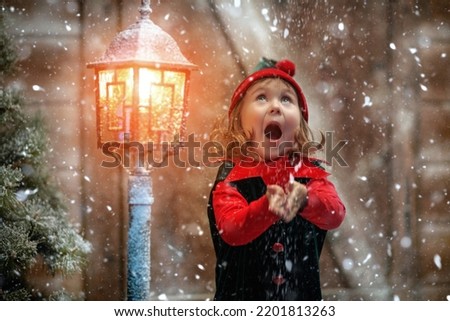 Winter Christmas tale. A funny cute elf girl catches snowflakes and rejoices, standing at a snow-covered wooden hut and lantern.